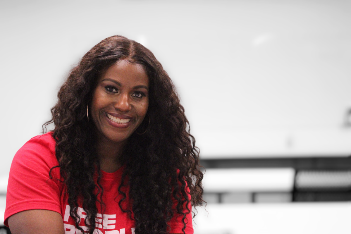 Portrait-style image of Stacy Davis Gates, sitting in the third row of a classroom, facing away from the front of the classroom. At the far wall is a blank whiteboard, out of focus. She is wearing a red t-shirt with the words "Free People Read Freely" in white lettering, and she is smiling broadly.