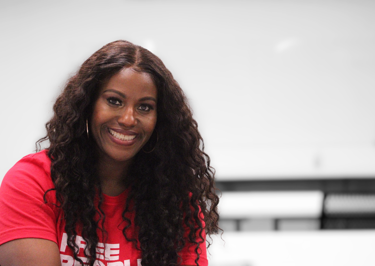 Portrait-style image of Stacy Davis Gates, sitting in the third row of a classroom, facing away from the front of the classroom. At the far wall is a blank whiteboard, out of focus. She is wearing a red t-shirt with the words "Free People Read Freely" in white lettering, and she is smiling broadly.