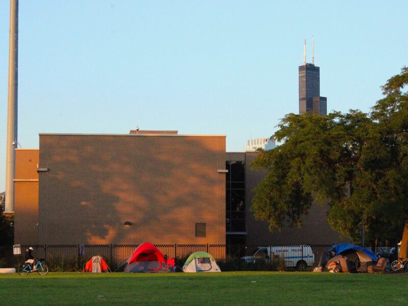 Brightly colored camping tents line the south wall outside the 12th District police station. A few asylum seekers are outside the tents; one is riding a blue Divvy bike across a wide, green field of grass in the foreground. The Sears/Willis Tower rises above the District on the right, where a large tree provides shade. A church steeple is in the background on the left. The sky is clear and blue.