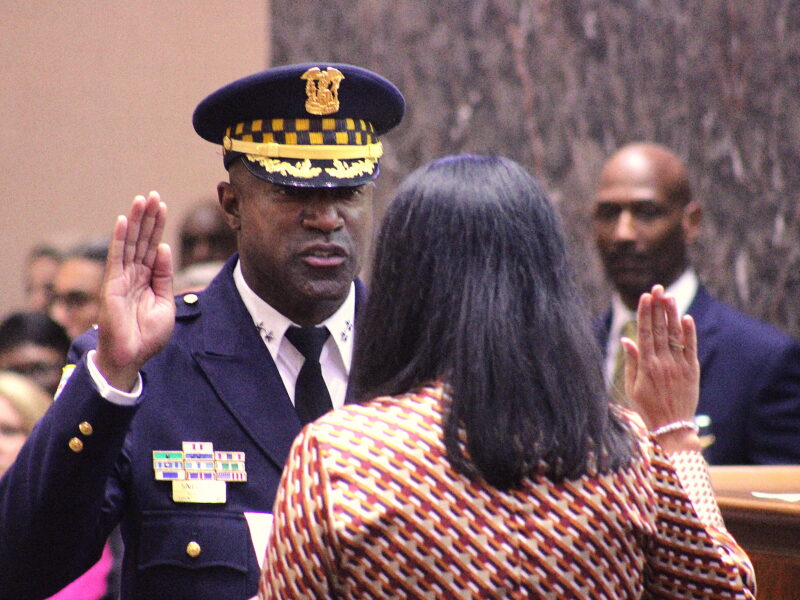 Larry Snelling, wearing the CPD's dress blue uniform, raises his right hand while facing the camera. Ana Valencia raises her right hand while facing Snelling. Valencia has long black hair and is wearing a multicolored red and black blouse. In the background, outgoing interim superintendent Fred Waller, a bald Black man wearing a suit, is slightly out of focus and facing the camera and Valencia, watching Snelling take the oath of office. The wall behind them is gray marble. Several other people are out of focus in the background.