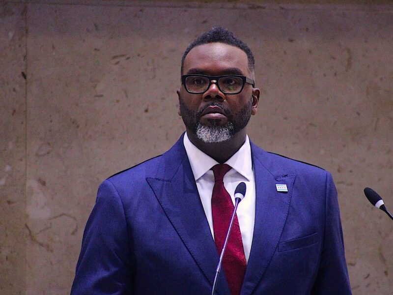Mayor Brandon Johnson, wearing glasses, a red tie and white shirt under a blue suit, stands in front of the gray marble wall in City Council chambers, two thin microphones in front of him.