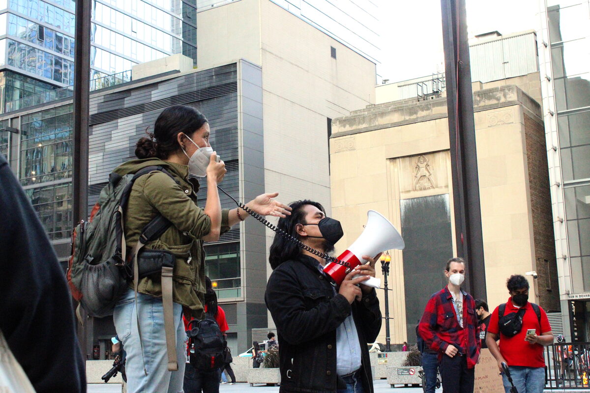A woman wearing an olive green jacket and jeans, as well as a surgical mask, speaks through the microphone of a bullhorn. A Latinx man with shoulder-length hair and a black jacket, also wearing a mask, holds the bullhorn. The base of the Picasso statue in Daley plaza is behind them, and the glass facade of the Federal Building is in the background.