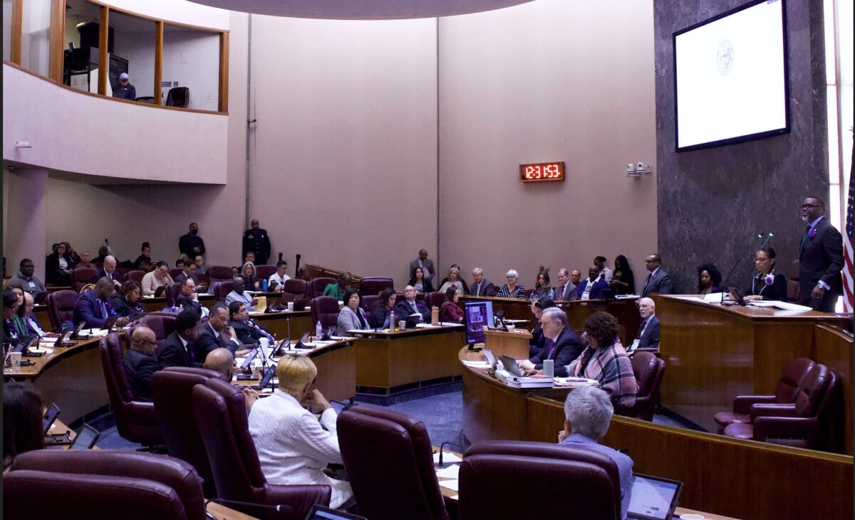 City Council chambers viewed from the side, with Mayor Brandon Johnson presiding from a dais on the right side of the picture and Council members arrayed at desks across the middle of the picture. A digital clock on the wall reads 12:31:52 in bright red digits, and a projector screen hung against a marble wall behind the dais displays the seal of the City of Chicago above and to the mayor's right.