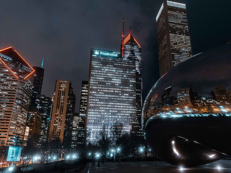 Glittering skyscrapers lit up against the night sky backdrop Cloud Gate, a shiny chrome bean-shaped sculpture in Millennium Park. The sculpture takes up the right half of the picture while buildings that line the north end of the park are seen in the background.